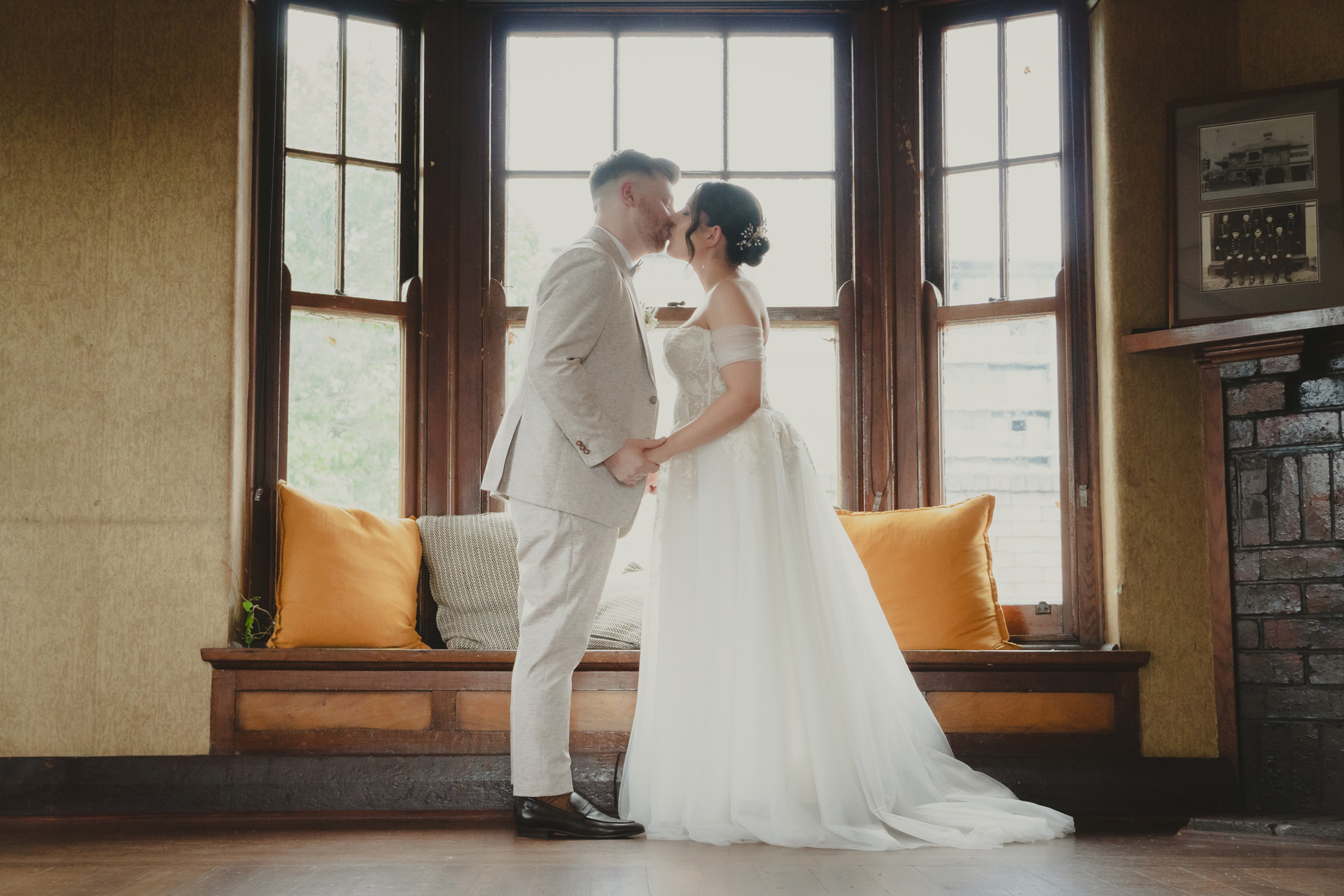 Bride in white gown and groom in beige suit holding hands and kissing by window seat with cushions.