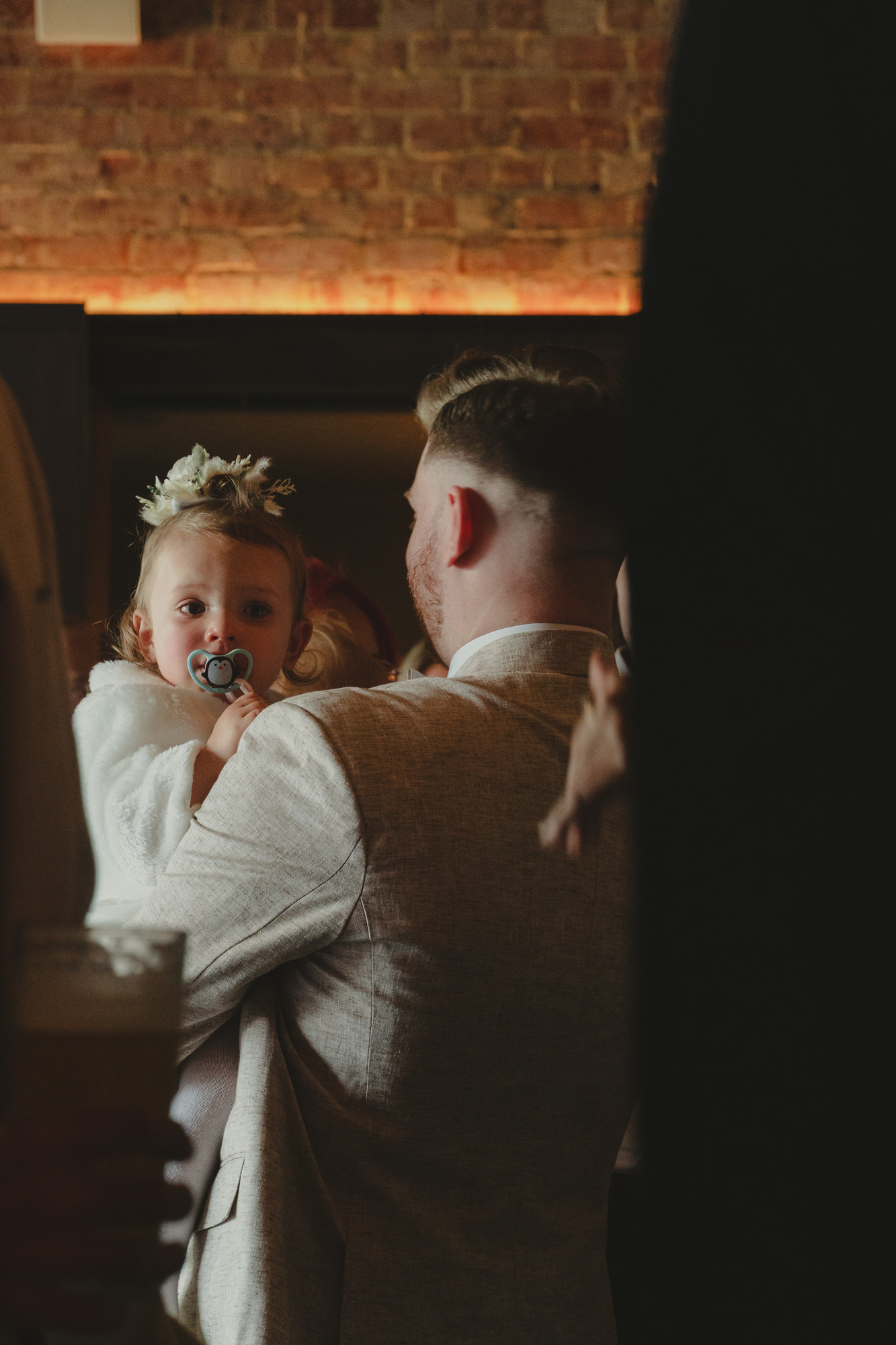 Man in beige suit holding a young child with a pacifier and floral headband indoors.