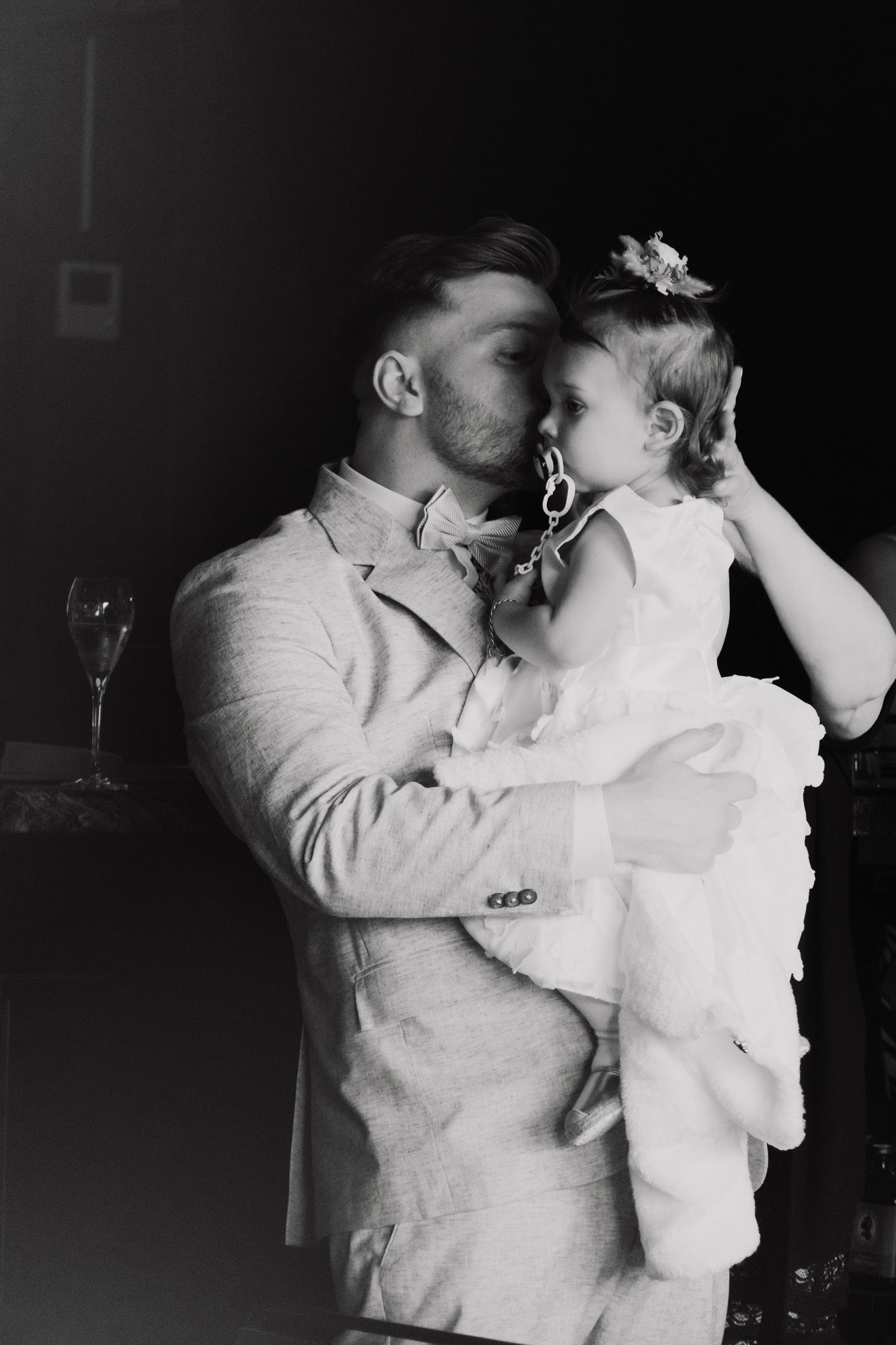 Man in suit gently kissing a young girl in a white dress holding a pacifier.