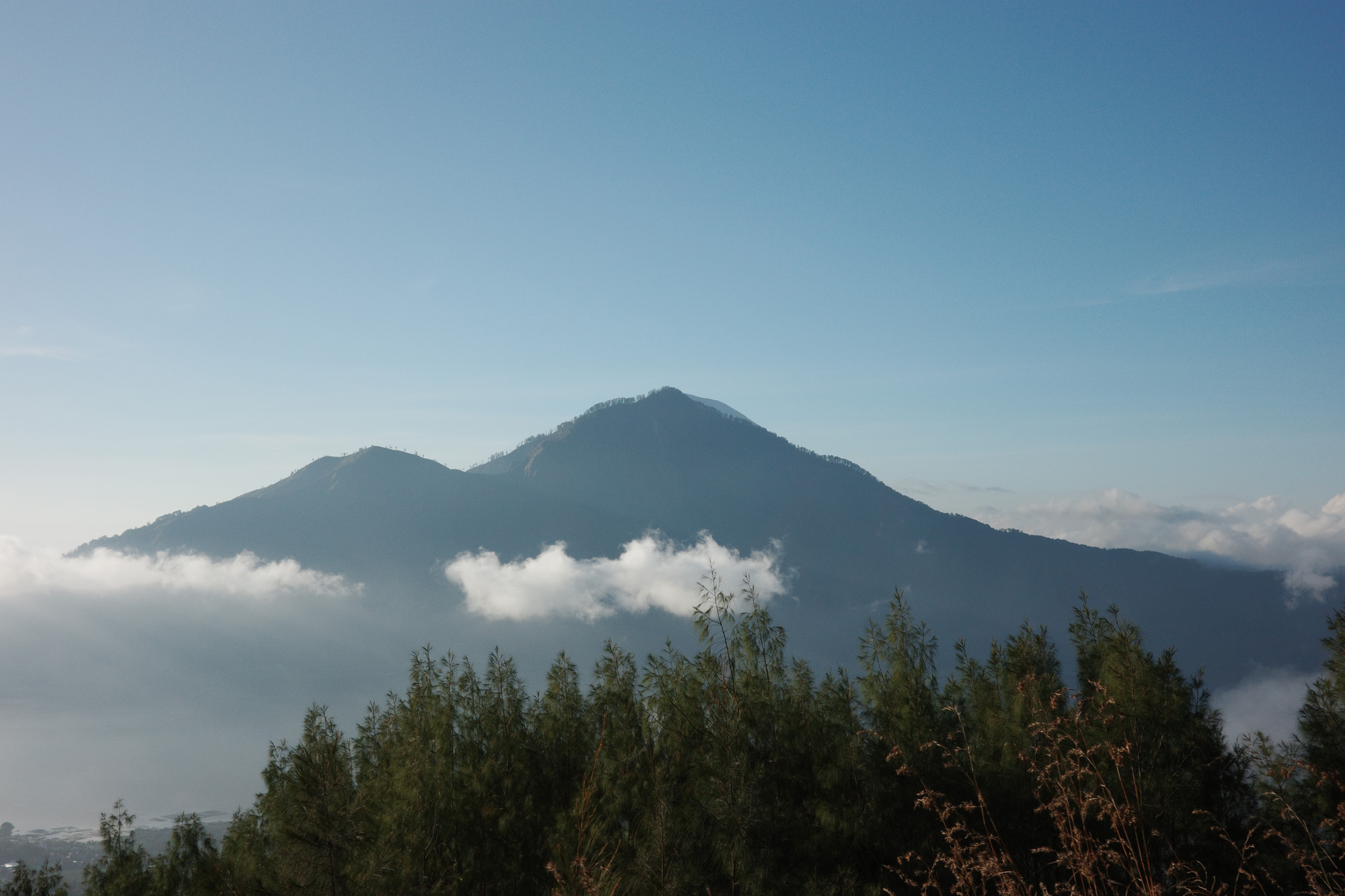 Mountain peaks rising above mist and clouds with trees in the foreground under a clear blue sky.