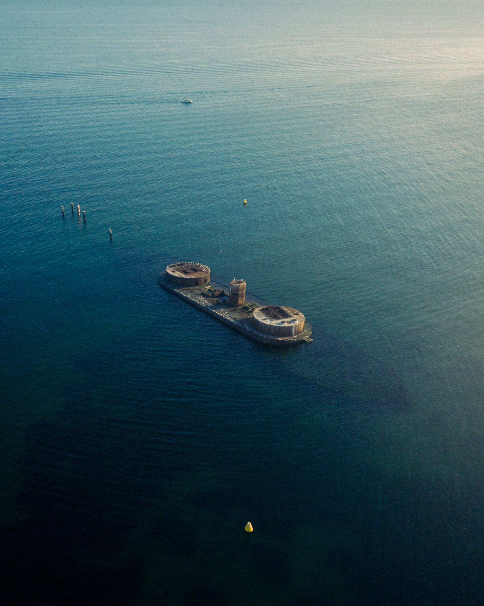 Aerial view of a large, abandoned concrete structure with two round towers in calm blue water.
