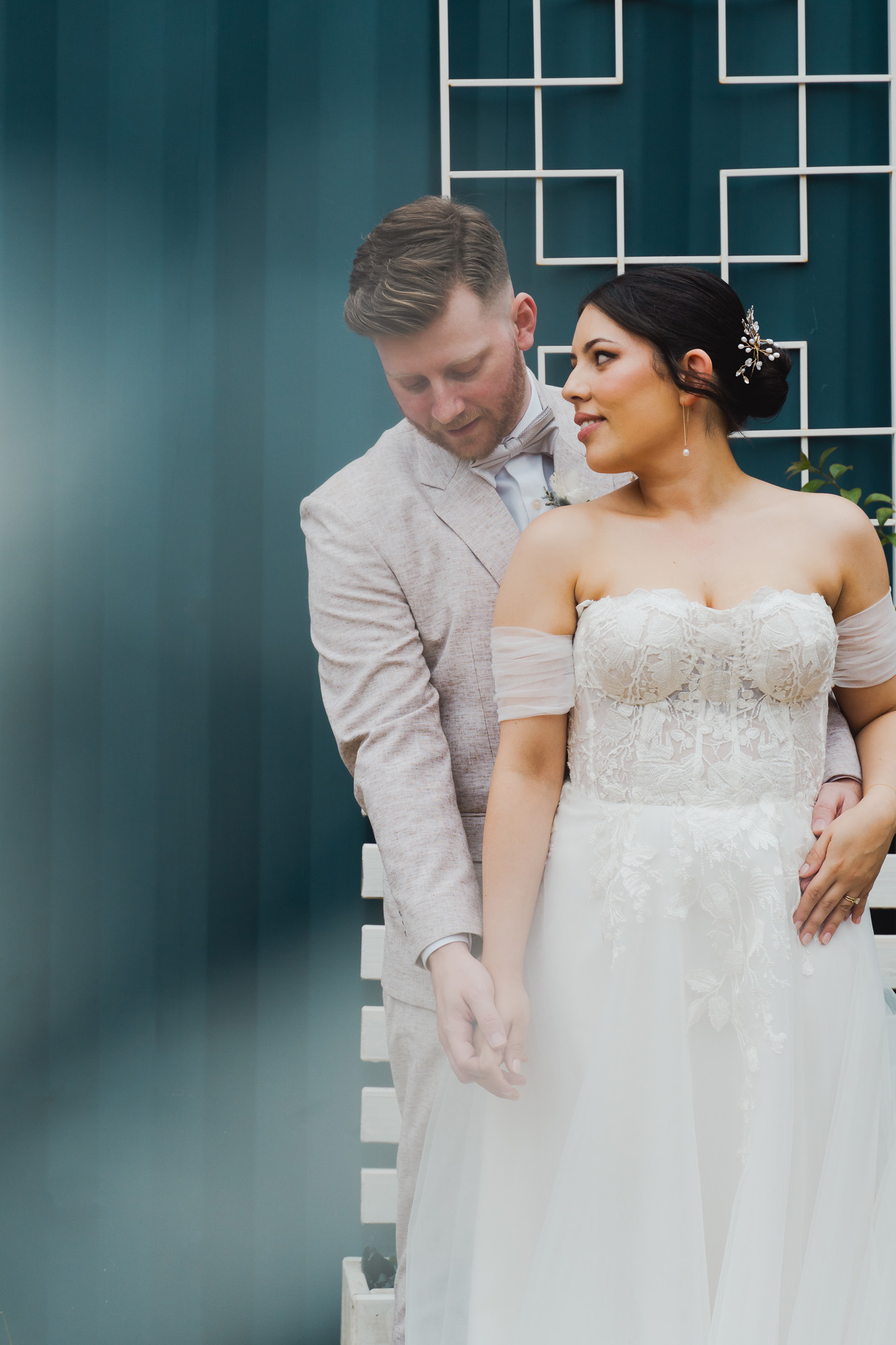 Bride in white lace dress and groom in light suit hold hands against a blue wall with a white grid.