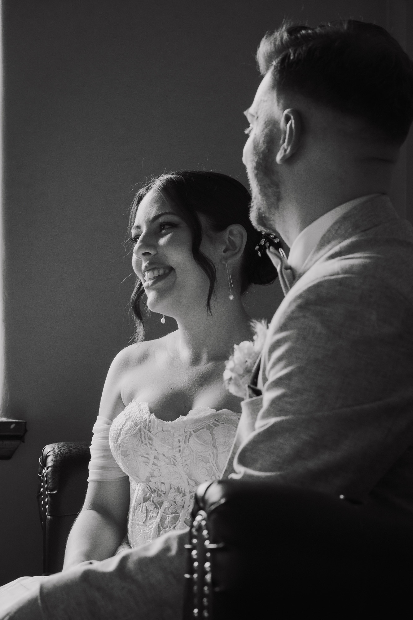 Black and white photo of a smiling bride in lace dress sitting beside a man in a suit indoors.