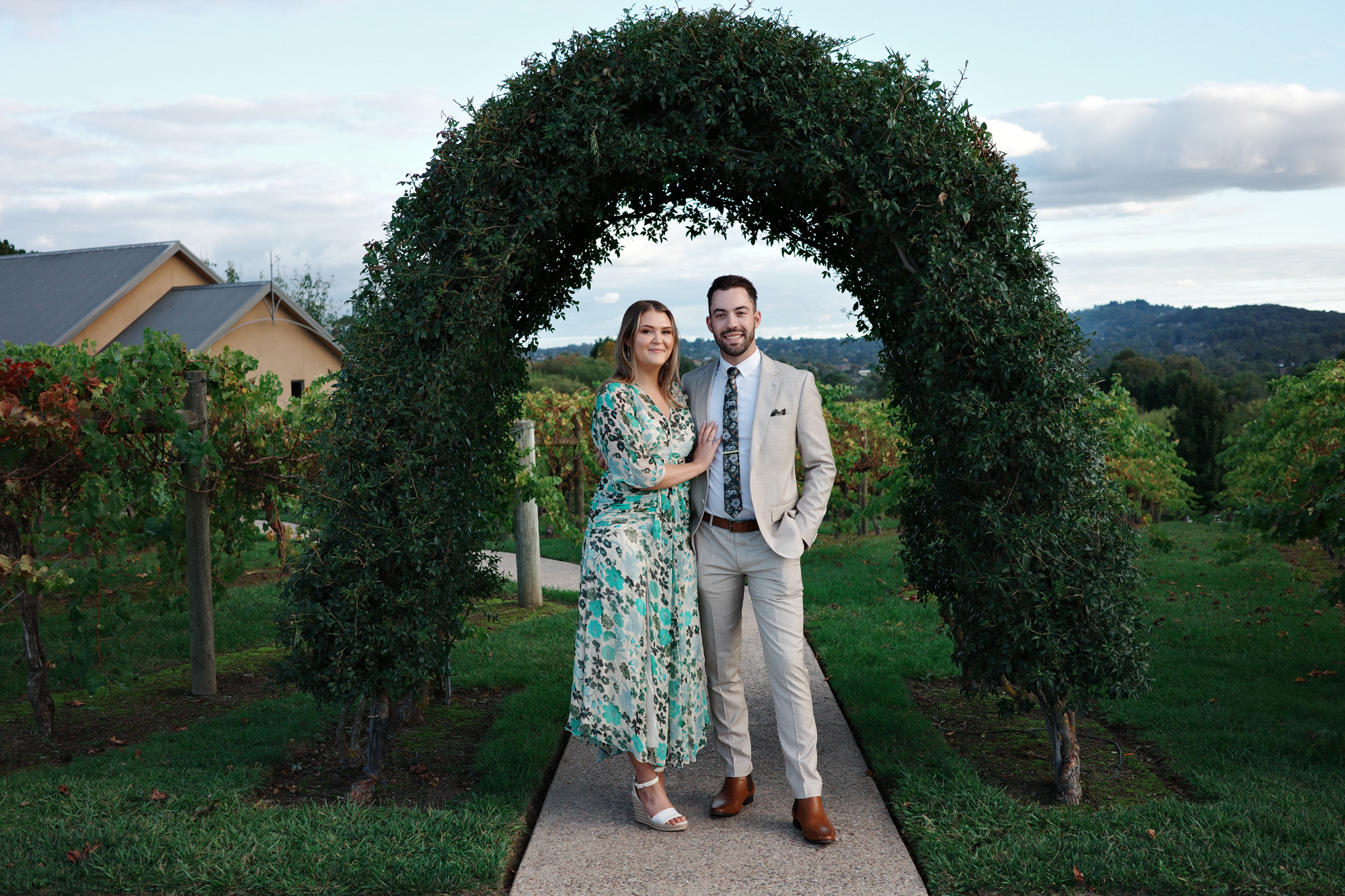 Couple posing under a leafy arch in a vineyard with hills and blue sky in the background.
