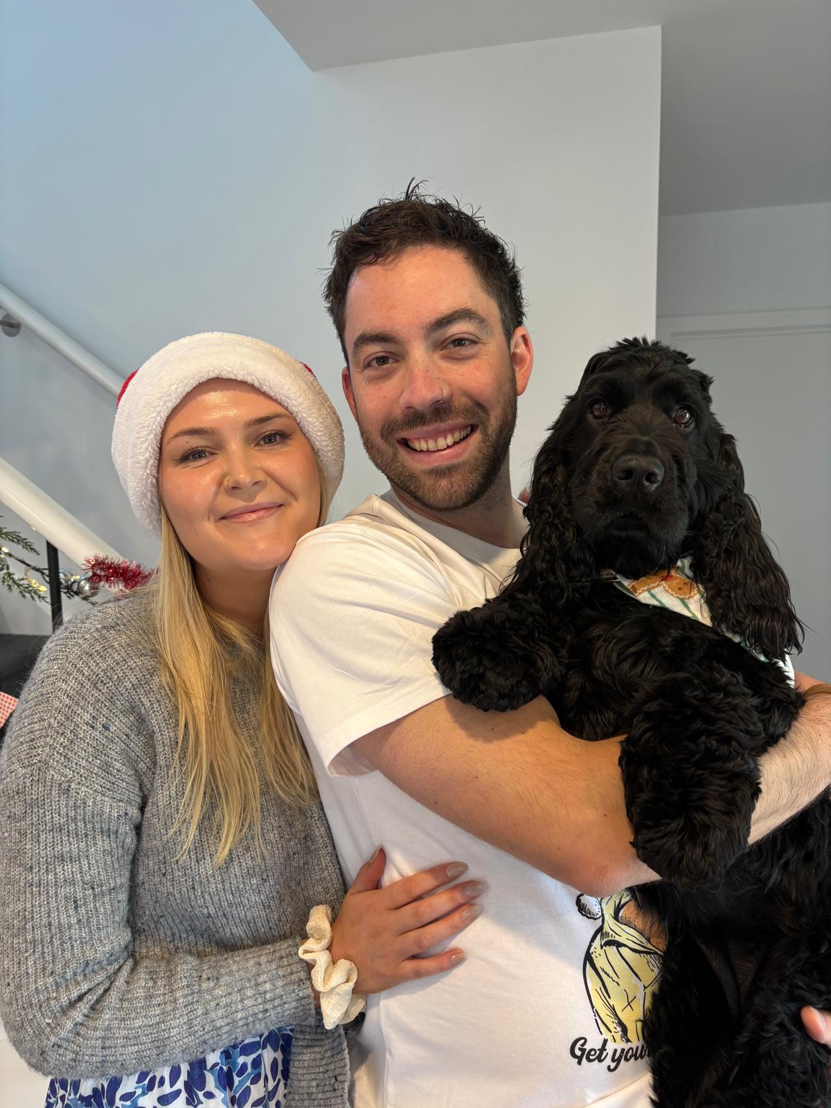Smiling couple indoors with woman in Santa hat and man holding a black dog wearing a bandana.