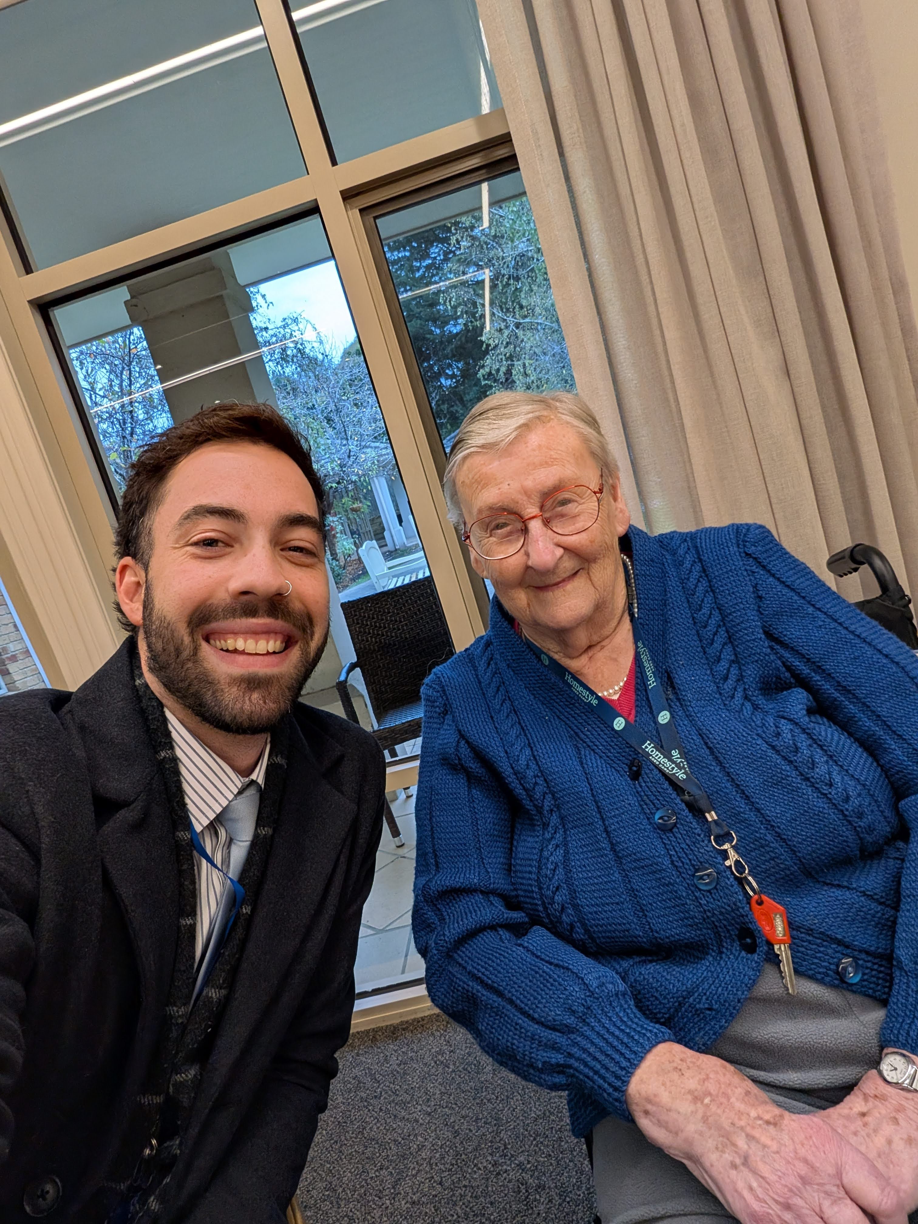 Smiling young man takes selfie with elderly woman in blue sweater sitting indoors near window.