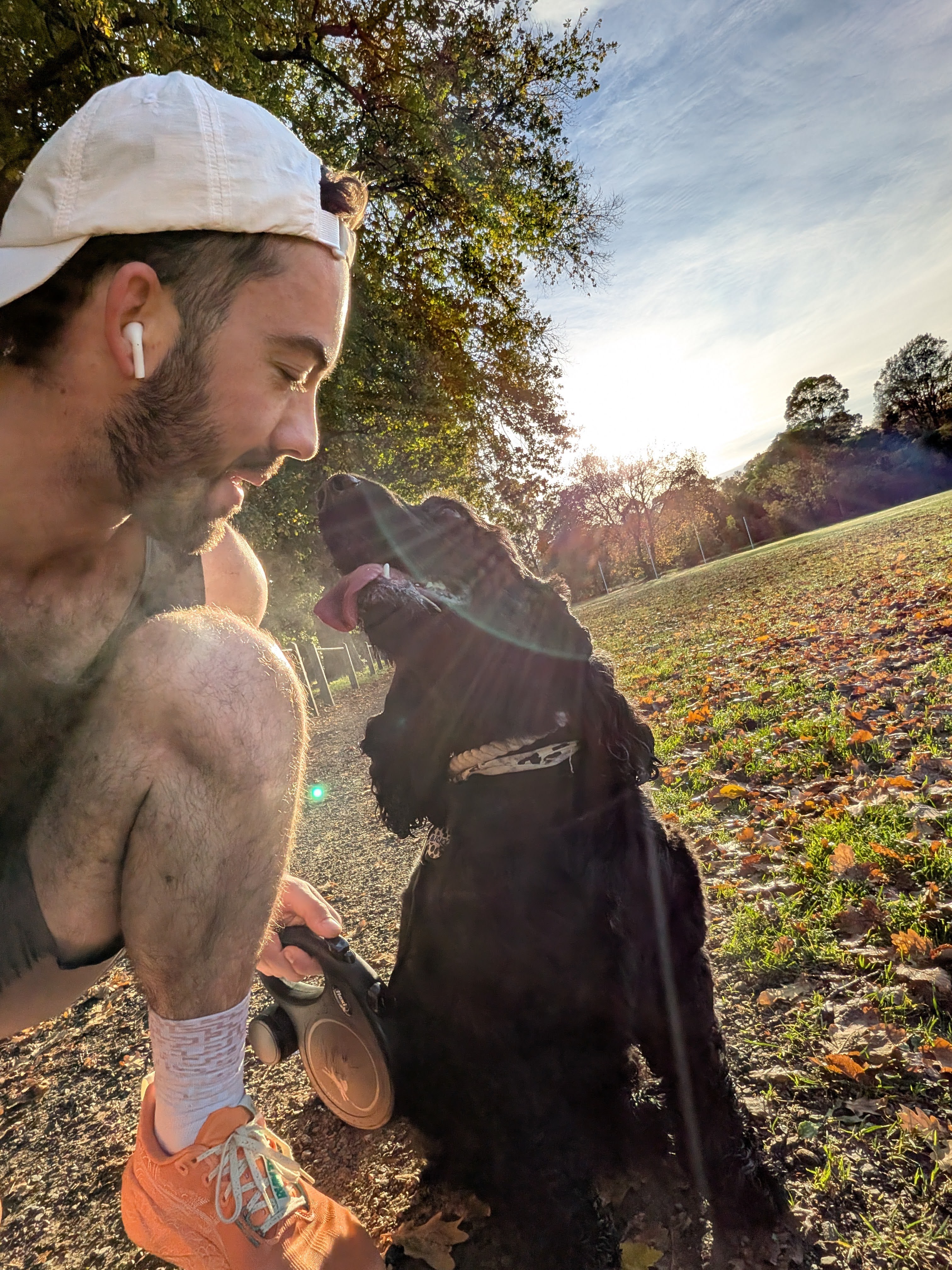 Man in white cap and orange shoes crouched, smiling at a black dog with tongue out in a sunlit park.