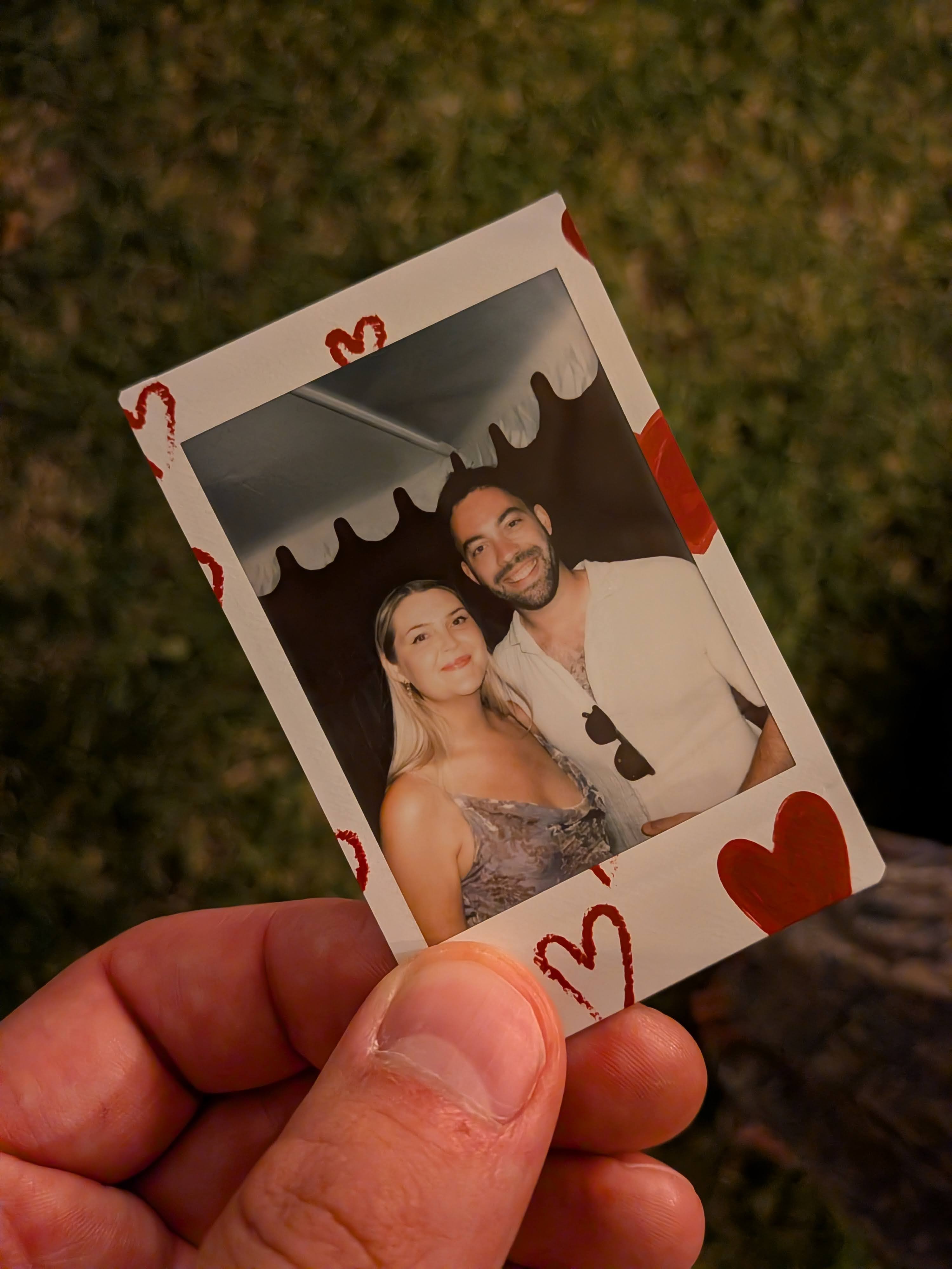 Hand holding a photo of smiling couple under a tent with heart decorations on the photo border.