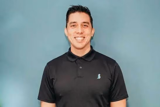 Smiling man with dark hair wearing a black polo shirt with a small blue logo on the chest, standing in front of a teal background.