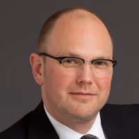 Portrait of a middle-aged man with a bald head and glasses wearing a black suit and tie against a dark gray background.