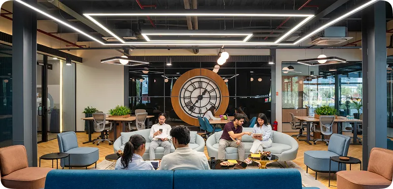 Modern office lounge with people seated on curved sofas, a large clock, and geometric ceiling lights.
