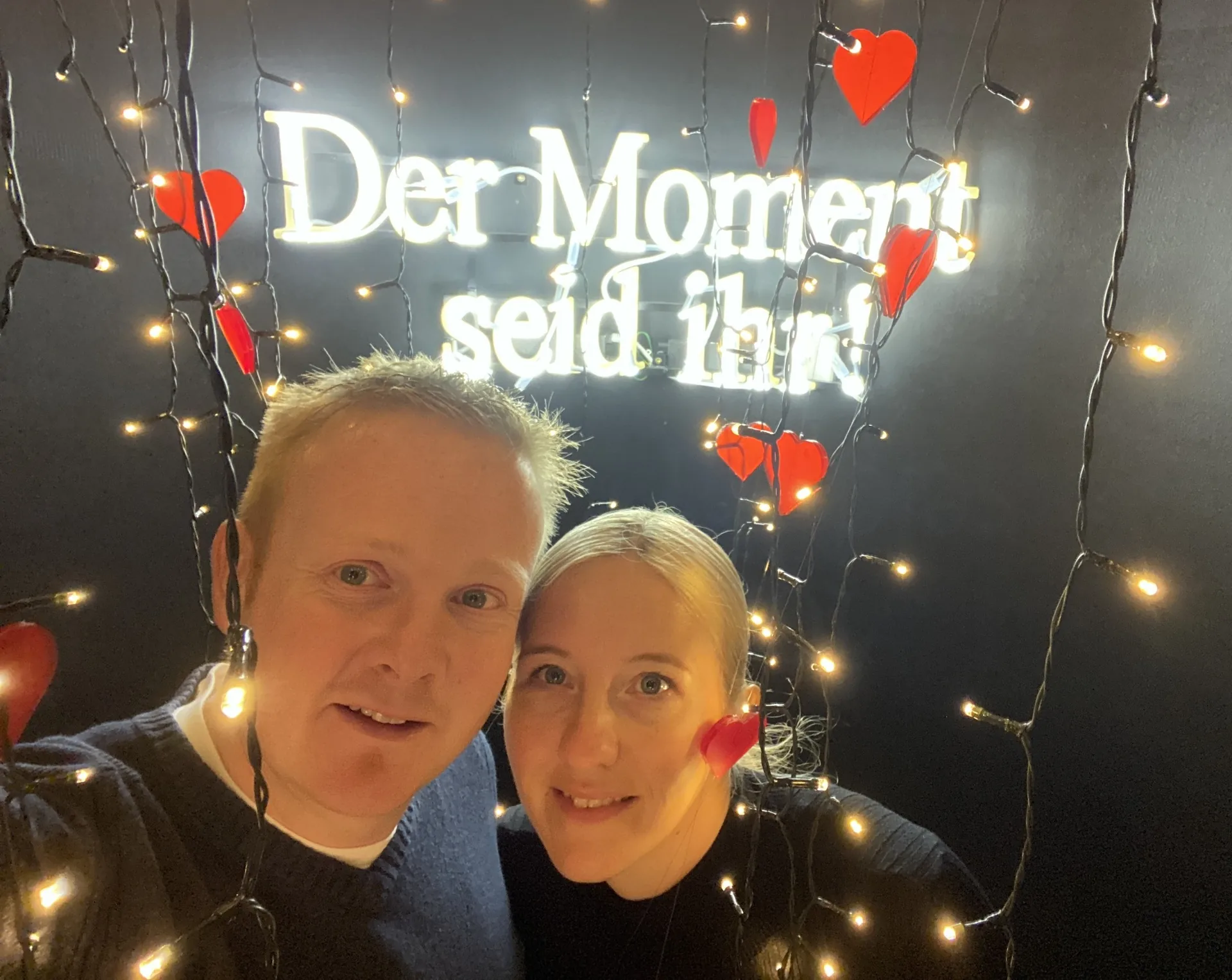 Denise und Thorben, die Gastgeber von Traumferienvilla, lächelnd vor einer Lichterkette mit der Aufschrift 'Der Moment seid ihr'