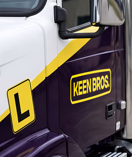 Close-up of a truck door with white and purple paint separated by a yellow stripe, featuring a yellow 'L' sign and a yellow-bordered 'KEEN BROS.' logo.