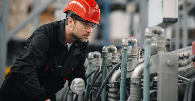 Man wearing red hard hat and safety glasses inspecting machinery with pipes and metal parts in an industrial setting.