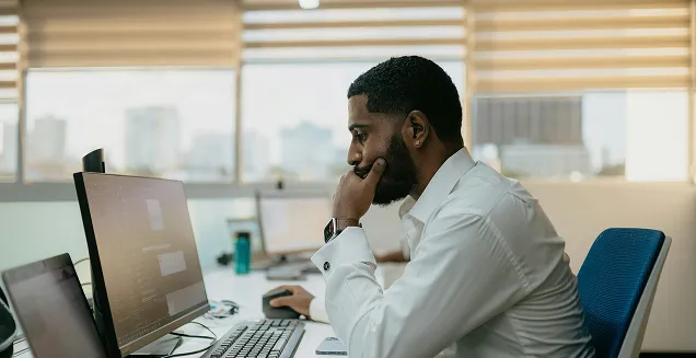 Man in white shirt sitting at a desk working intently on two computer monitors in a bright office with large windows.