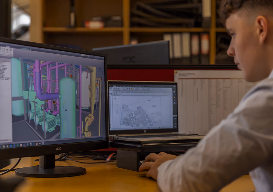 Young person working at a desk on a computer with dual screens displaying a 3D model of industrial piping and a site plan.