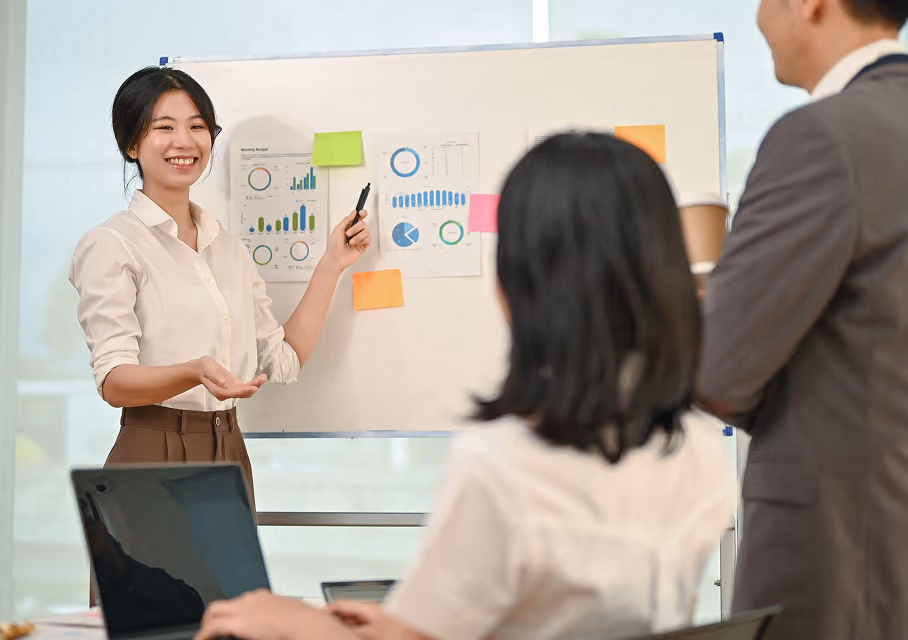 A woman in a white shirt presents charts and graphs on a whiteboard to two colleagues, one holding a coffee cup, in a bright office setting.