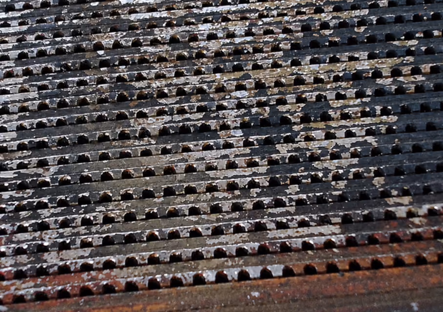 Close-up of a rusty, weathered metal grate with evenly spaced semicircular holes, showing chipped paint and varying shades of black, brown, and white.
