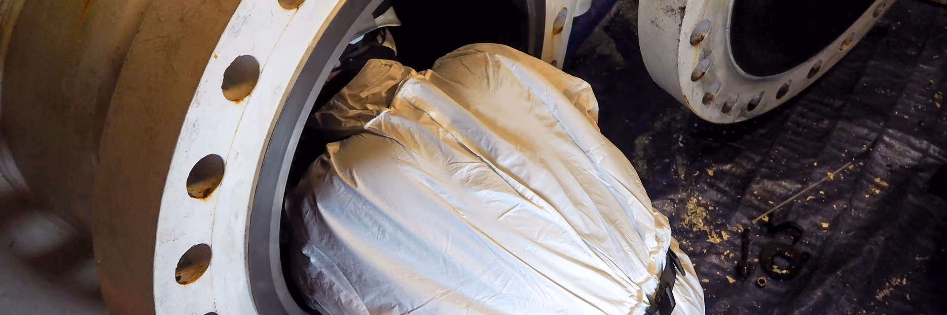 Person in a white protective suit working inside a large metal pipe flange in an industrial setting.