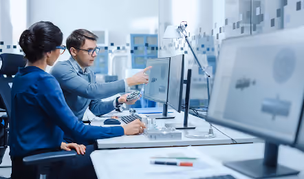 Two engineers collaborating at a desk in an office, one pointing at a computer screen displaying 3D mechanical parts while holding a component.
