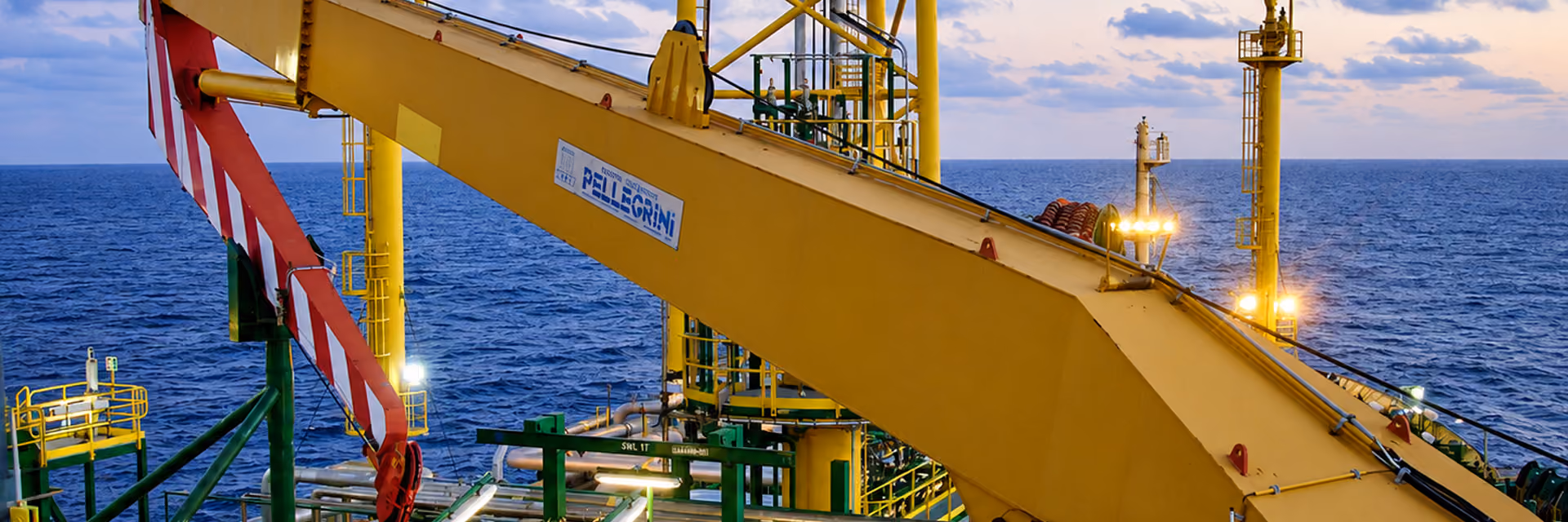 Yellow crane arm on an offshore oil platform with pipes and lights, set against a calm ocean under a cloudy sunset sky.
