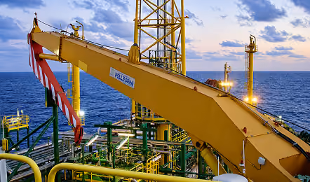 Yellow crane arm on an offshore oil platform with pipes and lights, set against a calm ocean under a cloudy sunset sky.