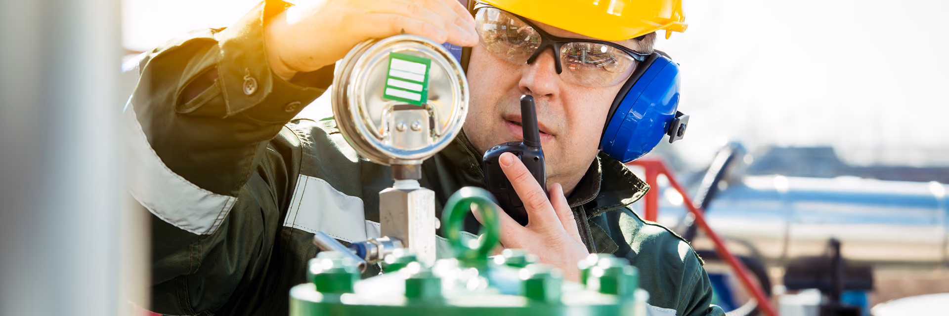 Industrial worker in a yellow hard hat and protective gear inspecting a gauge and speaking into a walkie-talkie at an outdoor facility.