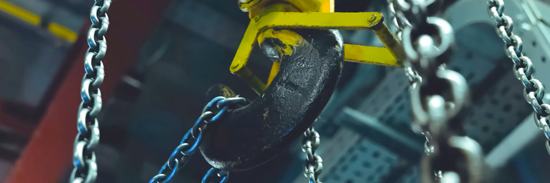 Close-up of a large black industrial crane hook attached to a yellow mechanism, surrounded by silver metal chains in a factory setting.