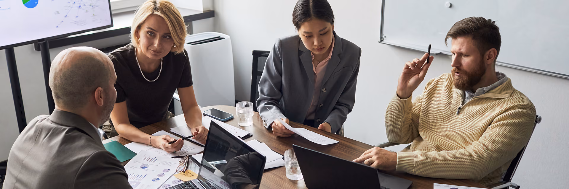 Four business professionals sit around a conference table with laptops and documents, engaged in a discussion in a modern office with a whiteboard and a large screen showing graphs and network data.