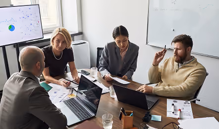 Four business professionals sit around a conference table with laptops and documents, engaged in a discussion in a modern office with a whiteboard and a large screen showing graphs and network data.