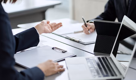 Two businesspeople in suits at a table with clipboards, a laptop, and a tablet; one person gestures with a hand while the other writes with a pen.
