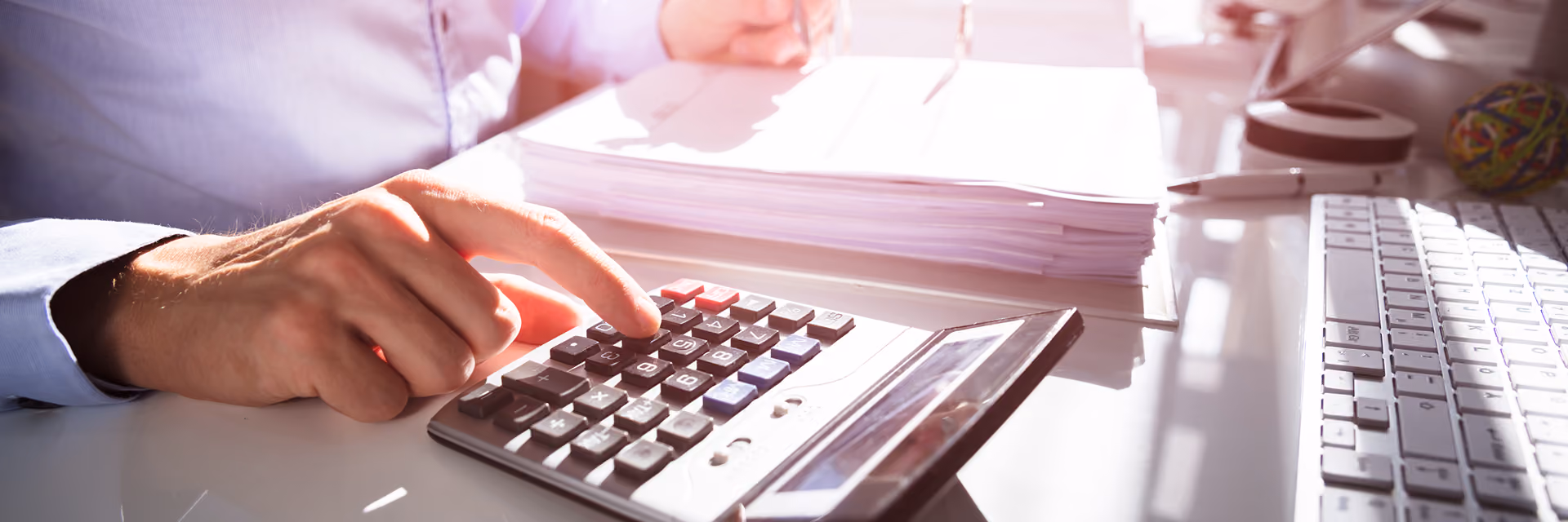 Person using a calculator with one hand while writing on documents at a desk with keyboard and tablet in a bright office setting.