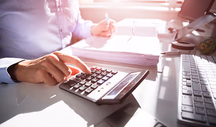 Person using a calculator with one hand while writing on documents at a desk with keyboard and tablet in a bright office setting.