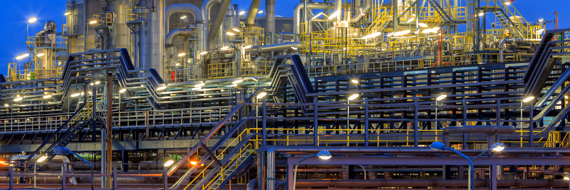 Industrial complex with multiple pipes, tall metal structures, staircases, and glowing lights against a twilight sky.