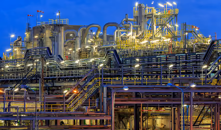 Industrial complex with multiple pipes, tall metal structures, staircases, and glowing lights against a twilight sky.