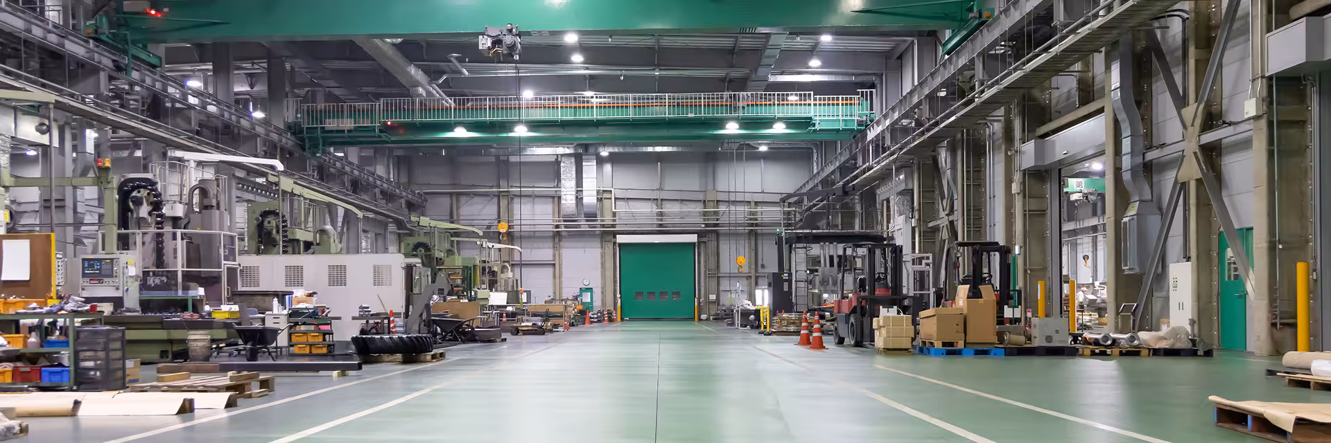 Interior of a large industrial warehouse with green floor, overhead cranes, stacked pallets, machinery on both sides, and bright ceiling lights.