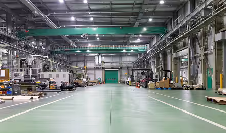 Interior of a large industrial warehouse with green floor, overhead cranes, stacked pallets, machinery on both sides, and bright ceiling lights.