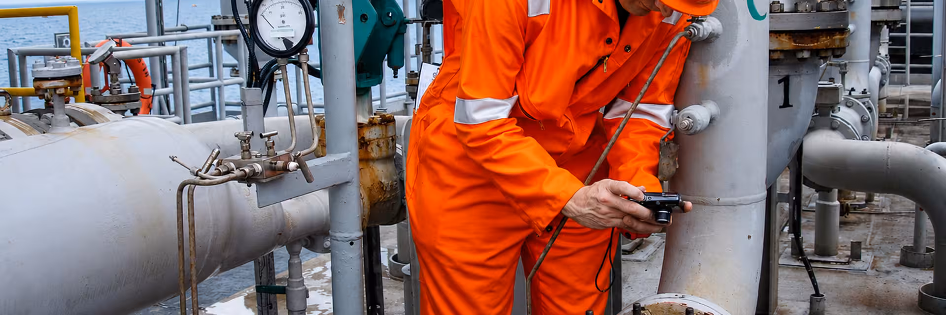 Worker in orange protective gear and helmet inspecting equipment and taking notes on an industrial platform with large pipes and gauges.