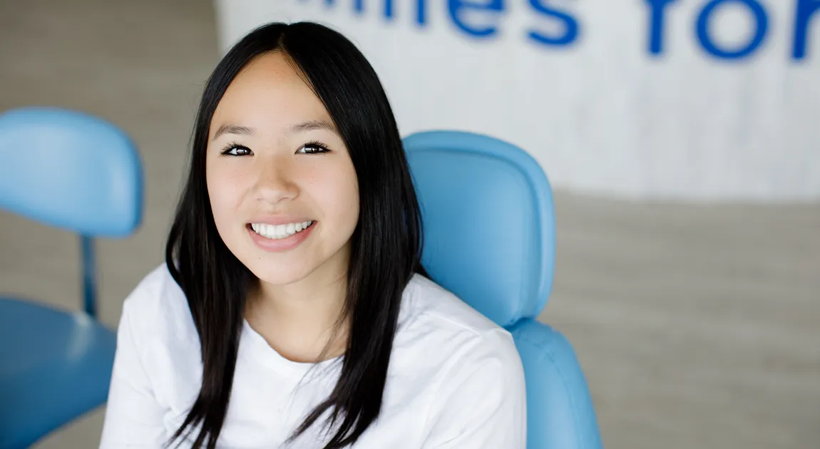 Smiling young woman with long dark hair sits in a blue office chair wearing a white shirt, creating a friendly, welcoming portrait.