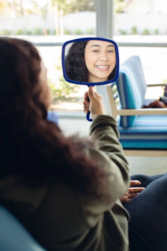 Inside a bus, a passenger holds a blue-framed hand mirror reflecting a smiling woman, creating a warm, playful moment between riders.