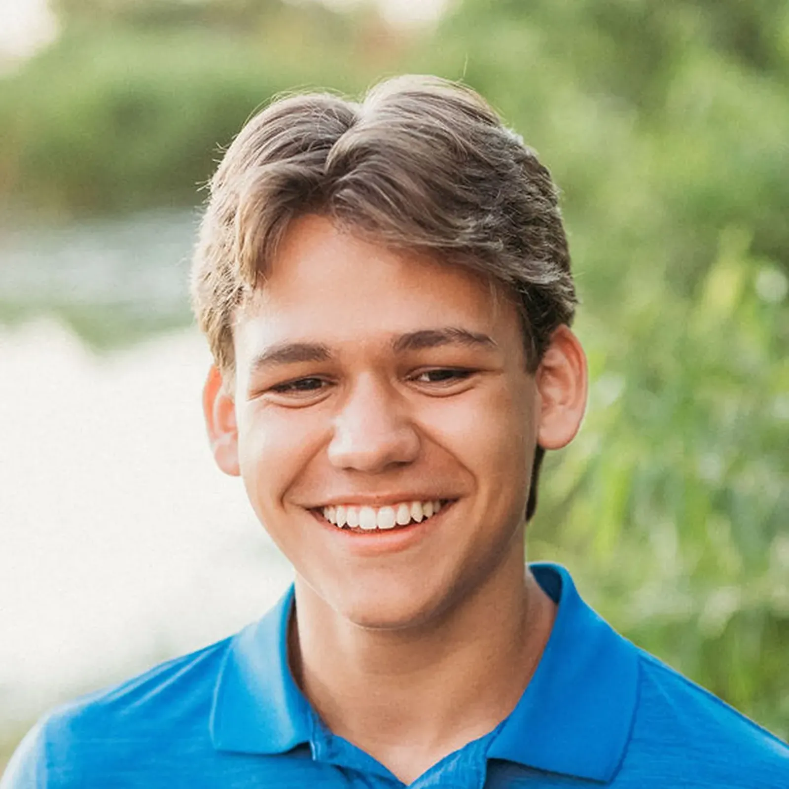 Smiling young man with brown hair wears a bright blue polo shirt outdoors, surrounded by soft green foliage and sunlight, conveying warmth.