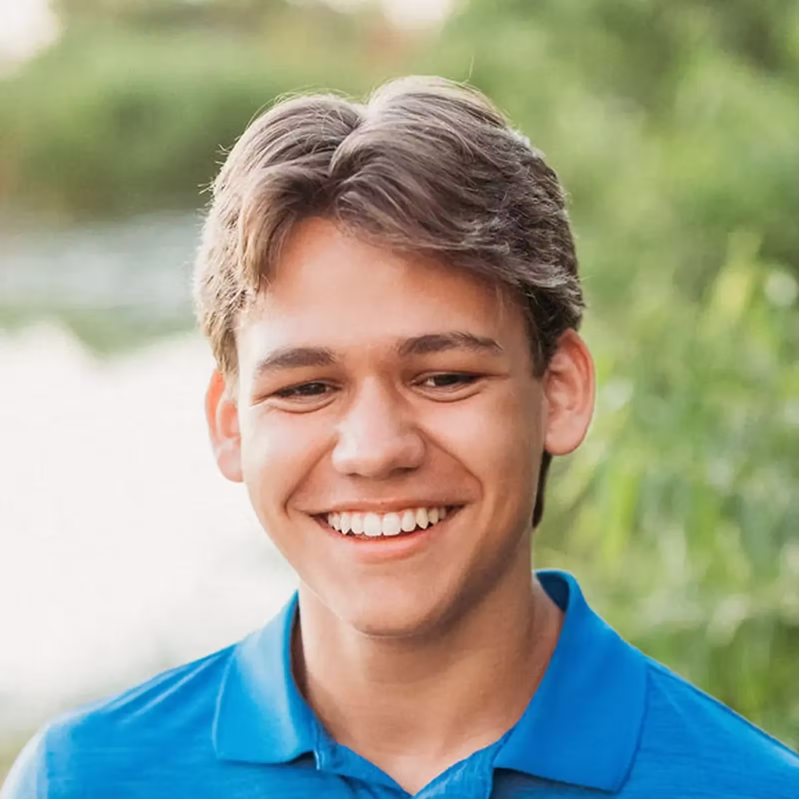 Smiling young man with brown hair wears a bright blue polo shirt outdoors, surrounded by soft green foliage and sunlight, conveying warmth.
