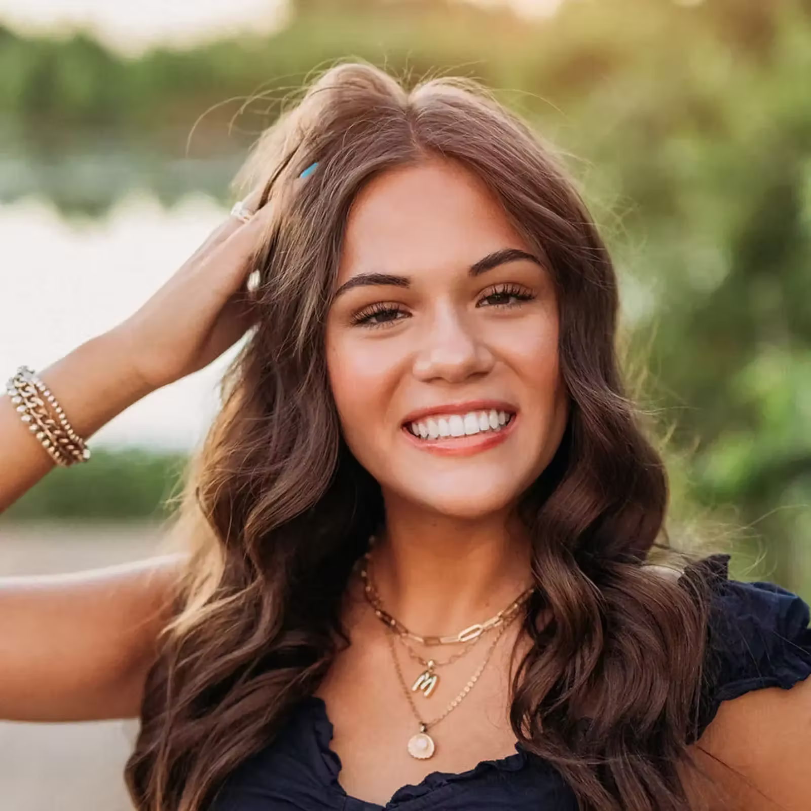 Smiling young woman with long, wavy brown hair poses outdoors by a lake, hand in her hair, wearing layered necklaces and a dark top.