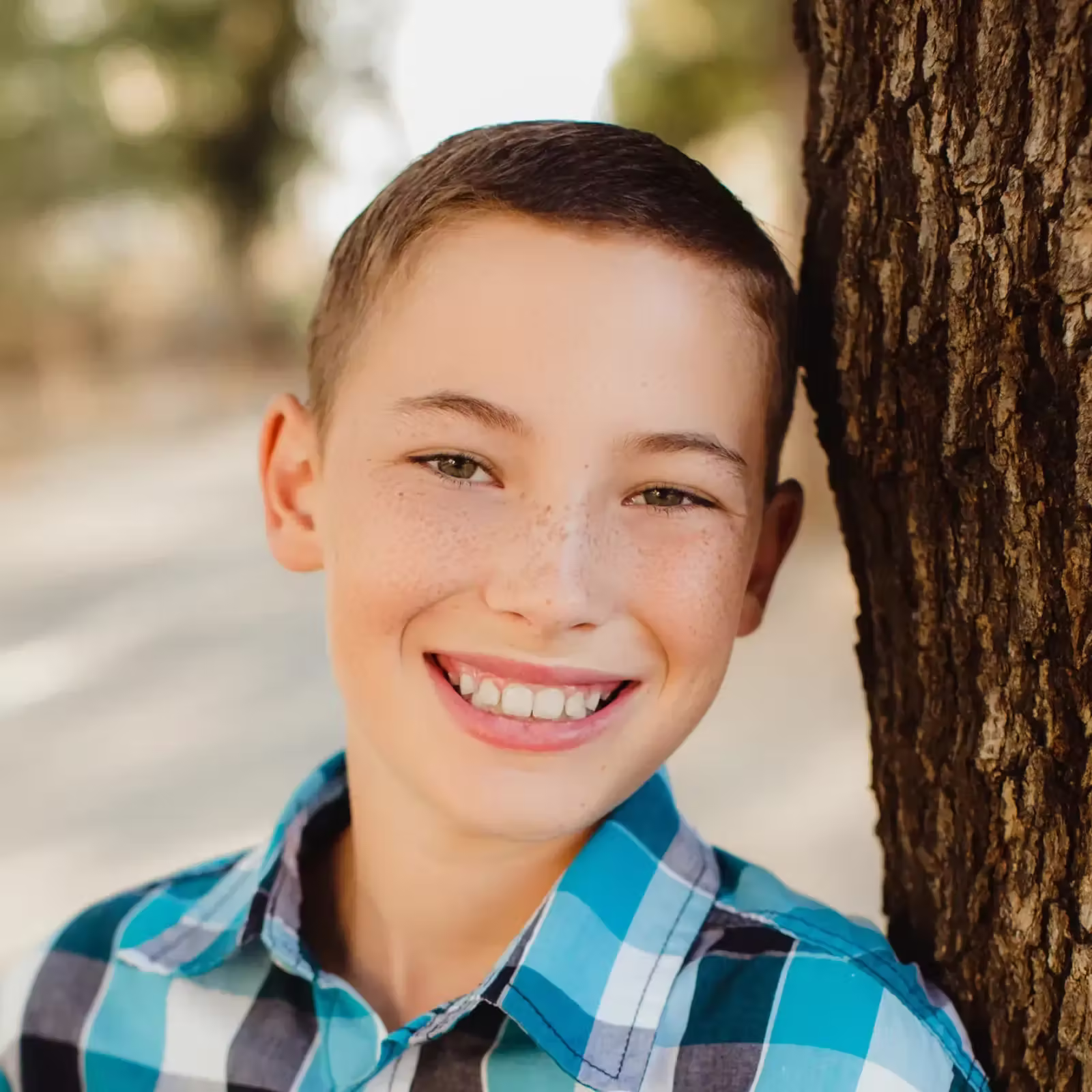 Smiling freckled boy in a blue plaid shirt leans against a tree outdoors, warmly lit by sunlight, conveying joy and carefree youth.