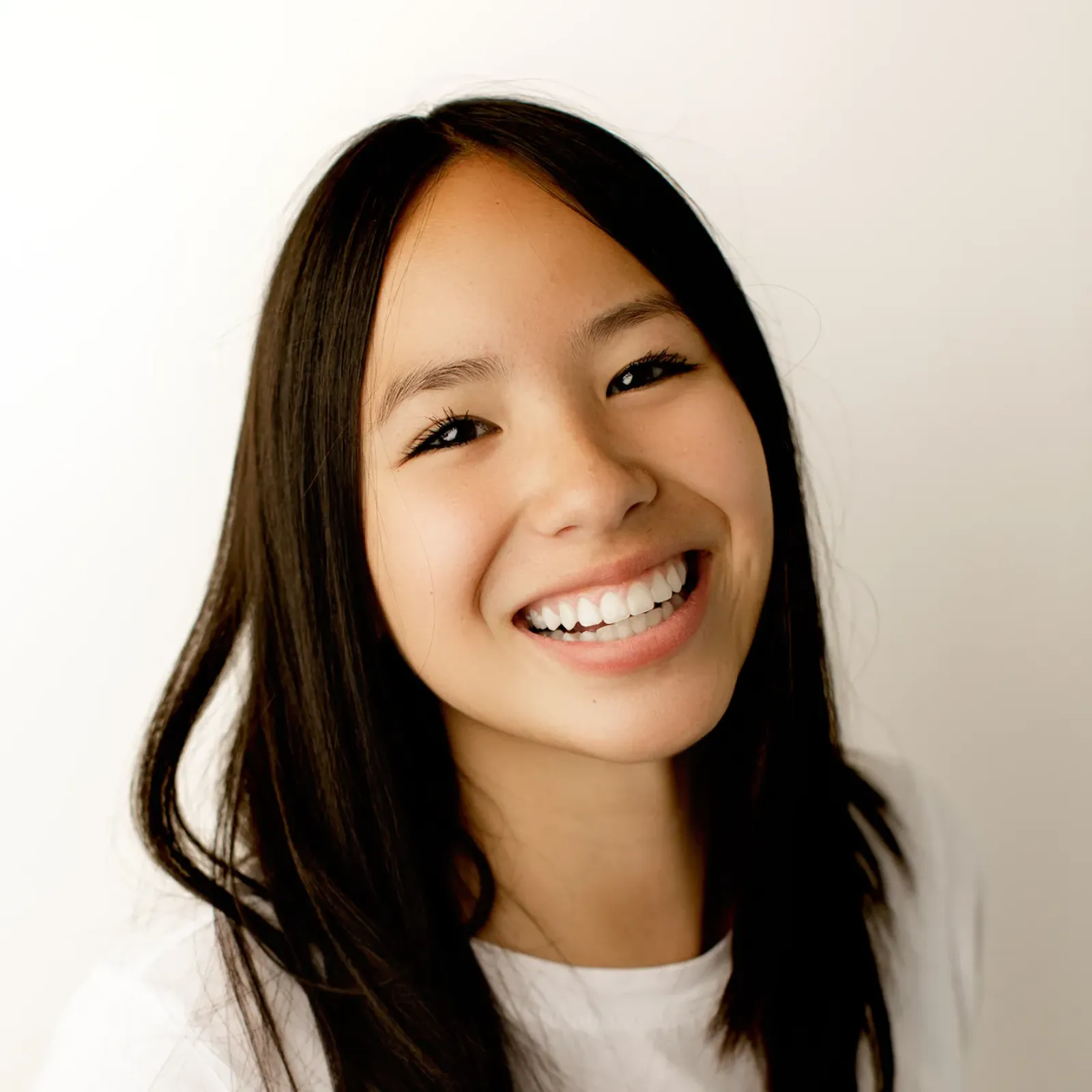 Close-up portrait of a smiling young woman with long dark hair in a white shirt, facing the camera against a light background; warm, joyful mood.