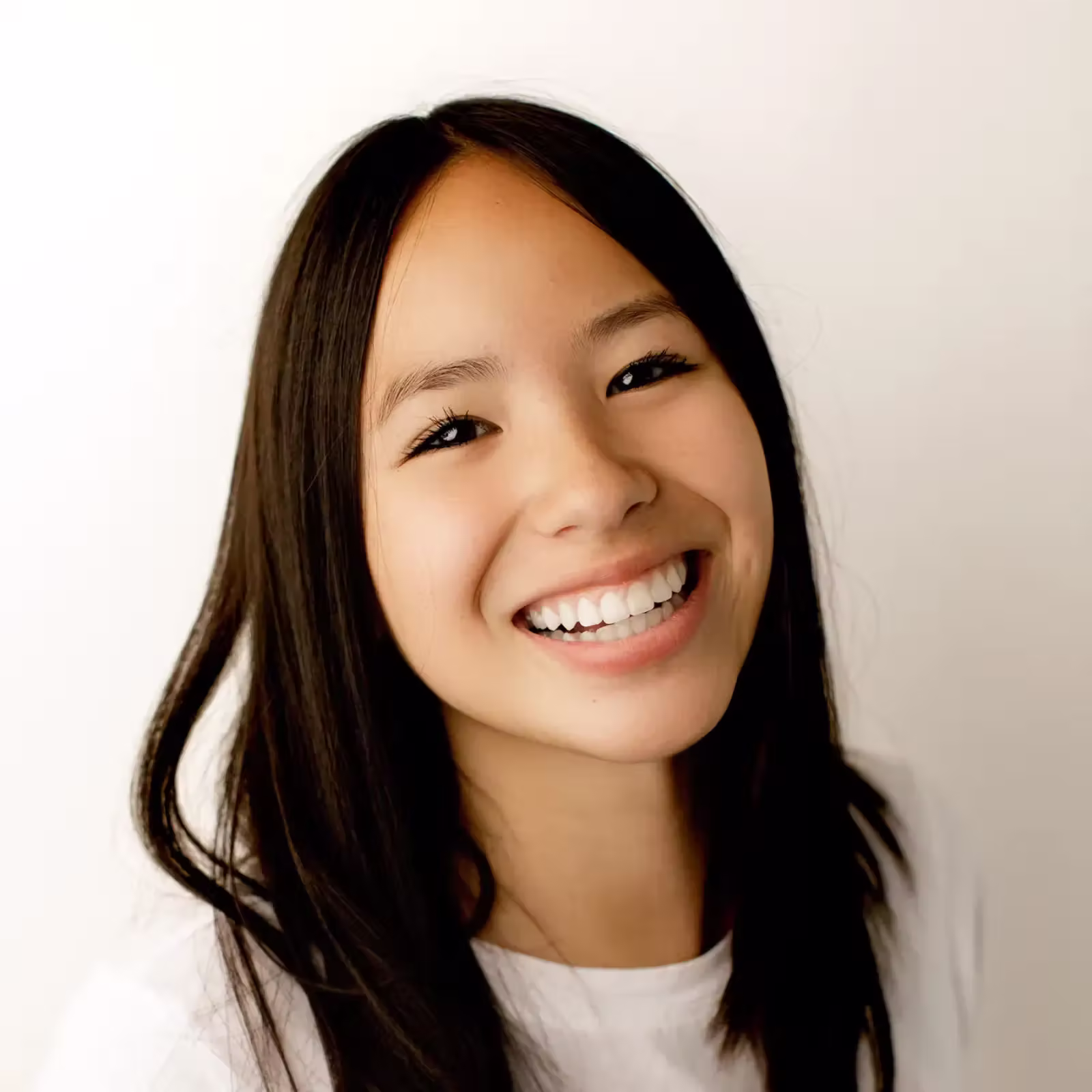 Close-up portrait of a smiling young woman with long dark hair in a white shirt, facing the camera against a light background; warm, joyful mood.