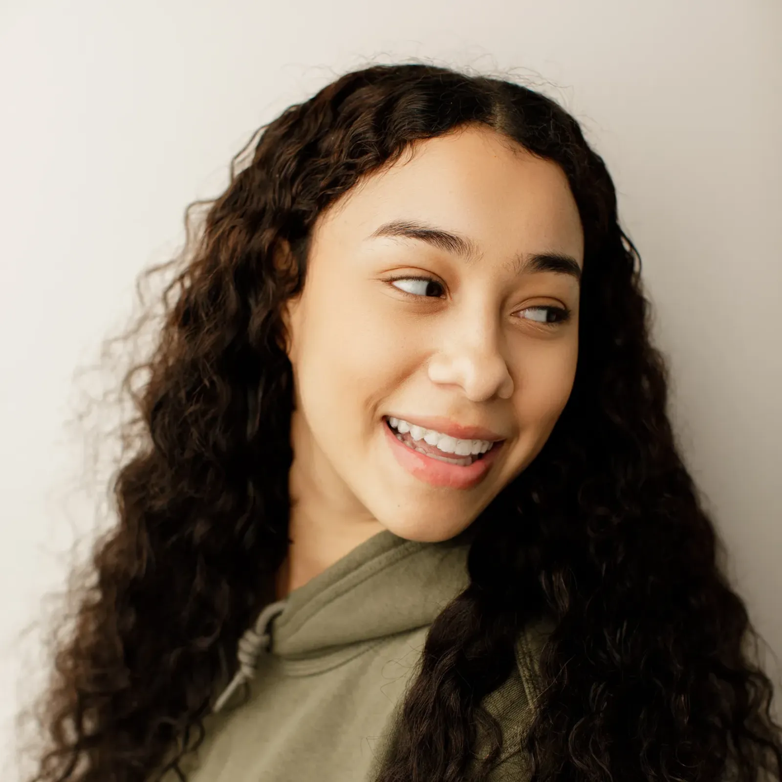Smiling young woman with long curly dark hair wears an olive-green hoodie, glancing to the side in a warm, friendly portrait.