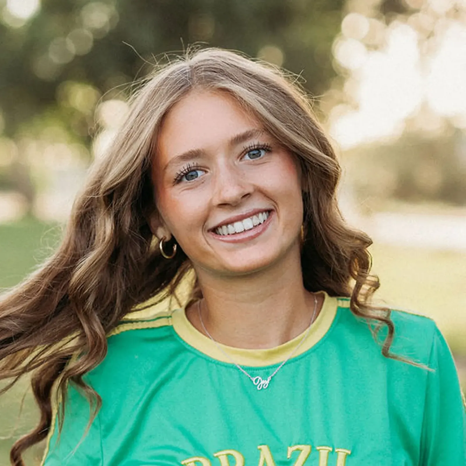 Smiling young woman with long wavy brown hair wears a bright green shirt outdoors, warmly lit by sunlight in a cheerful portrait.