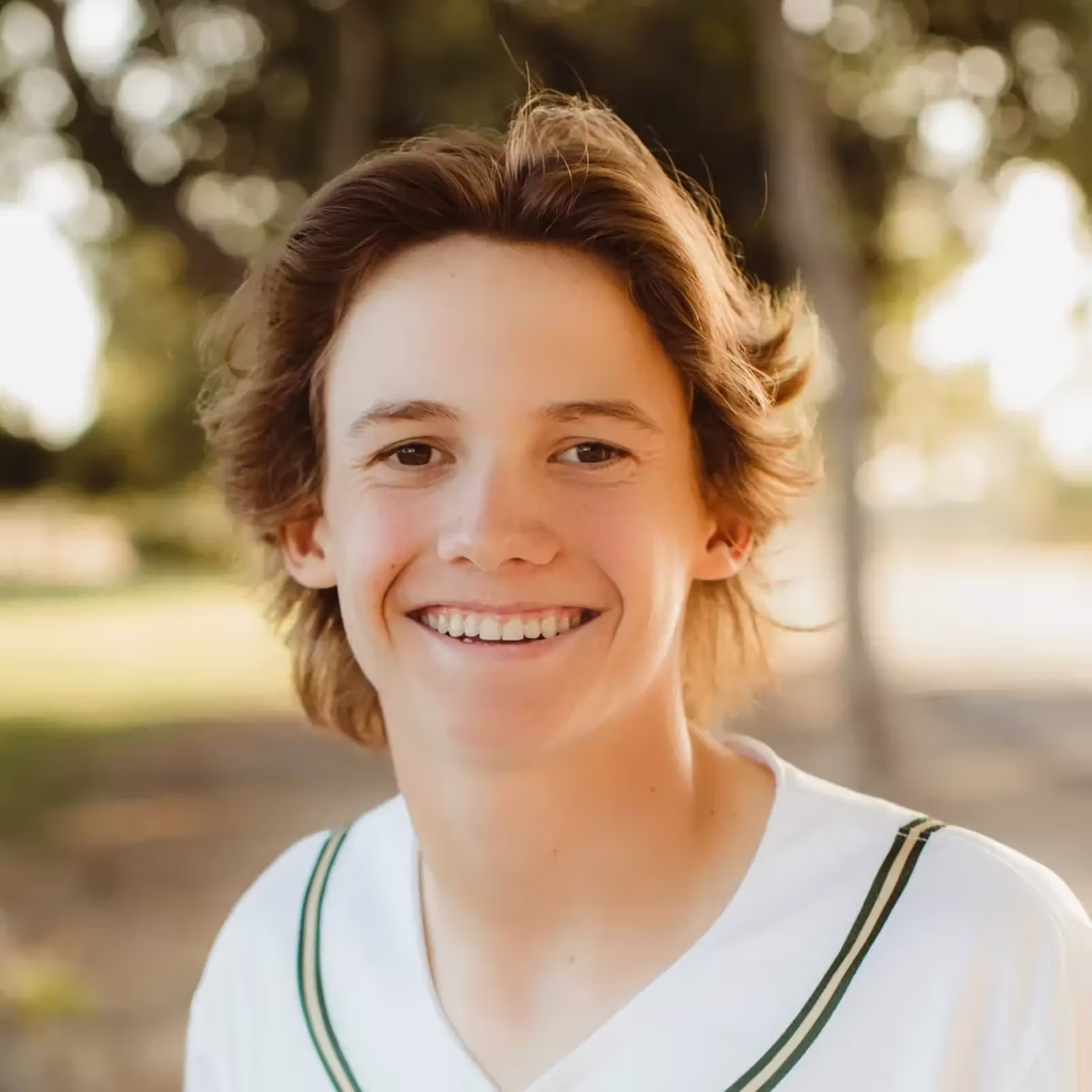 Smiling teenage boy with tousled brown hair wears a white sports jersey outdoors, warmly lit by sunlight in a park; cheerful, friendly mood.