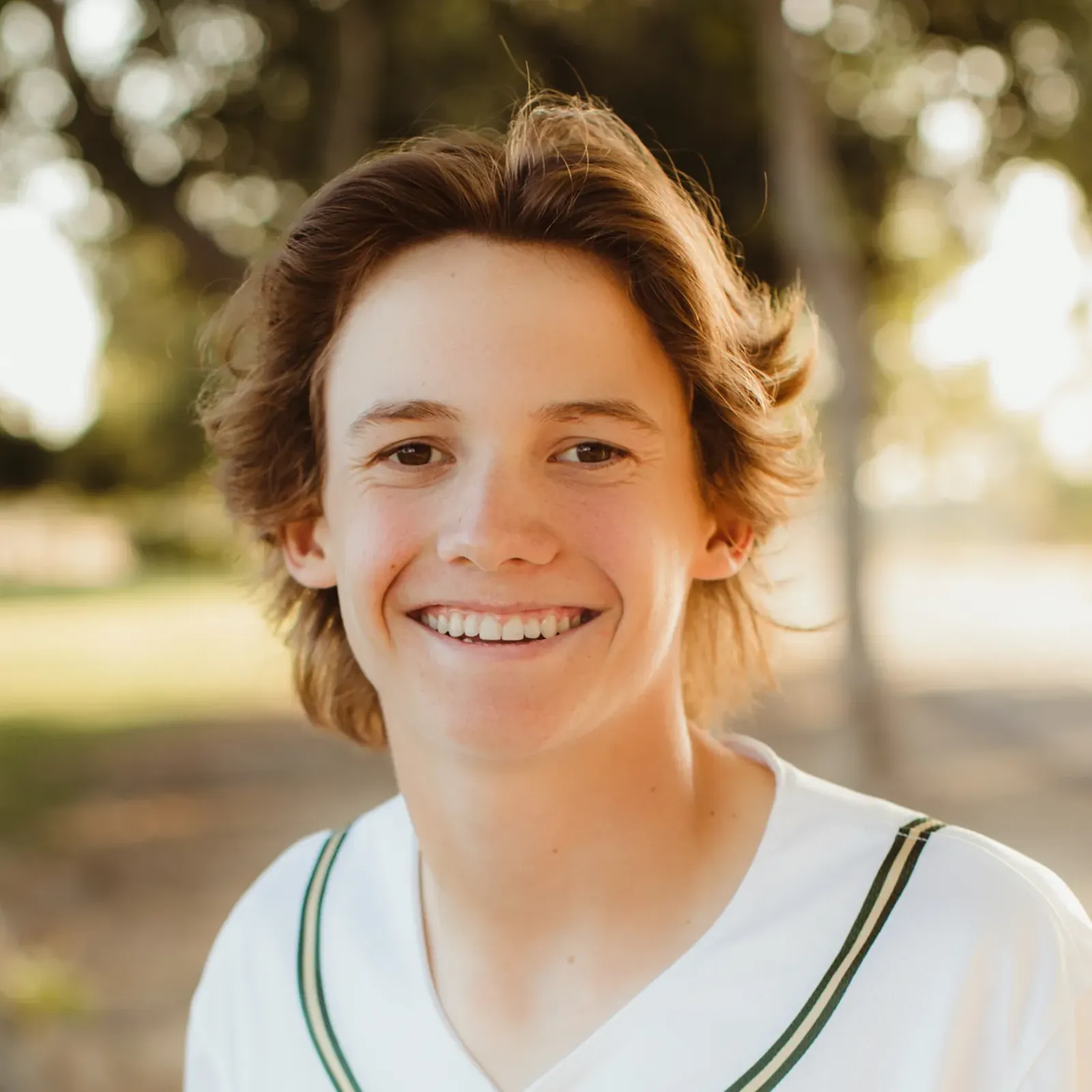 Smiling teenage boy with tousled brown hair wears a white sports jersey outdoors, warmly lit by sunlight in a park; cheerful, friendly mood.